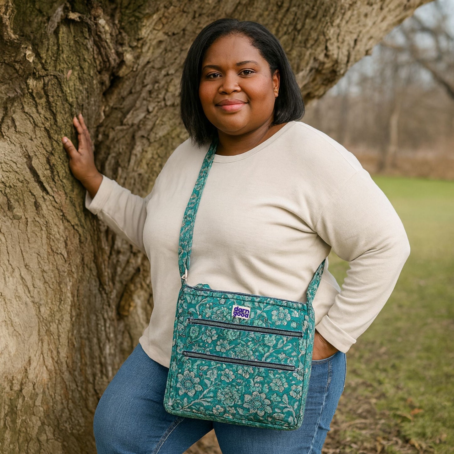 Woman standing outdoors with a green floral-patterned bag, leaning against a tree.