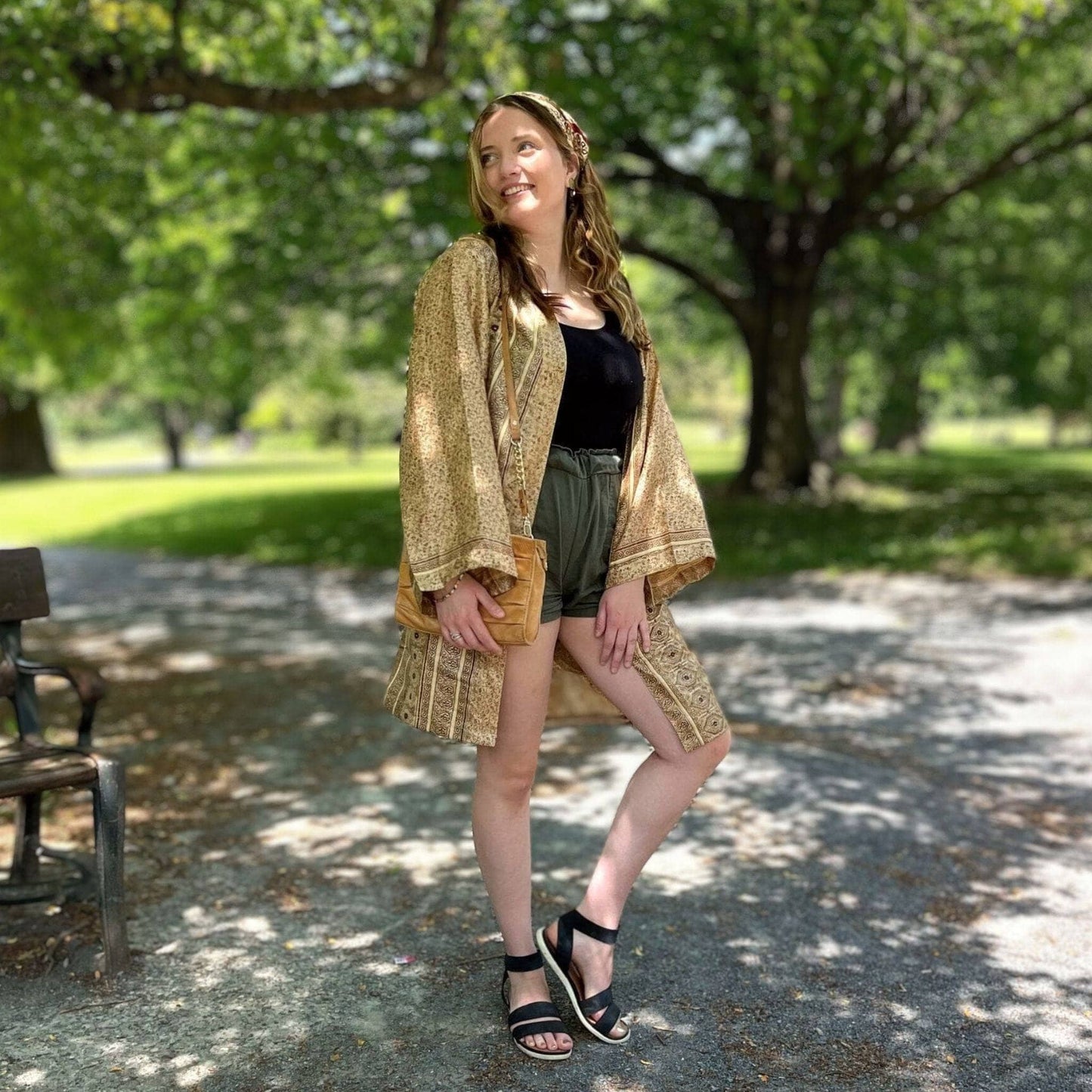 Woman in a gold kimono standing in a park with trees and a bench in the background.