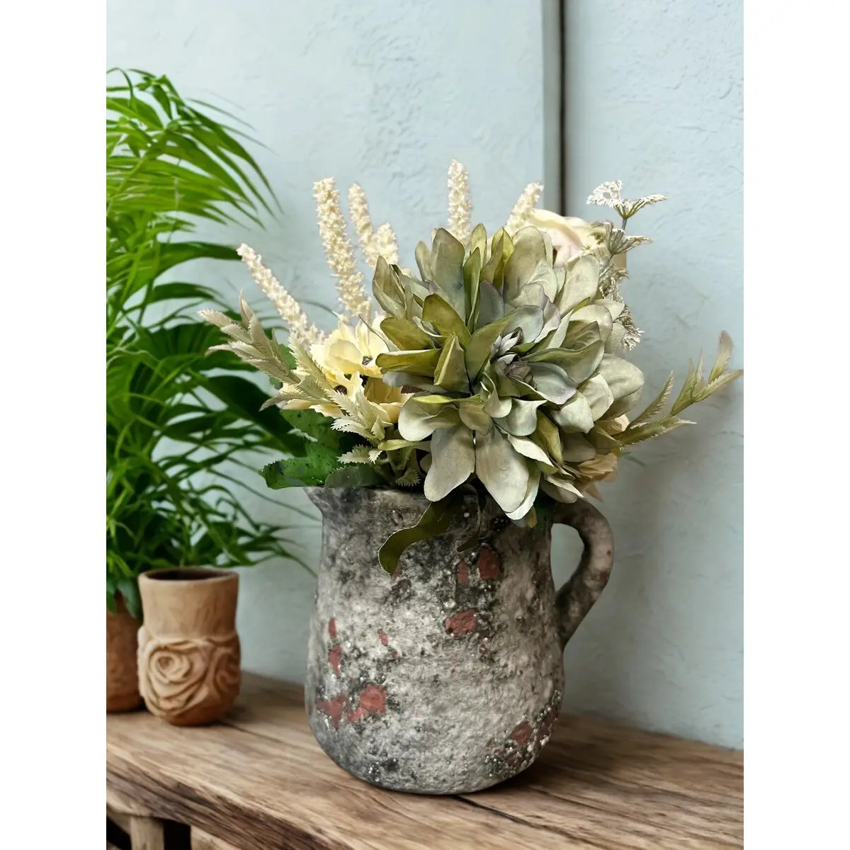 Decorative pitcher with artificial flowers on a wooden surface against a light blue wall.