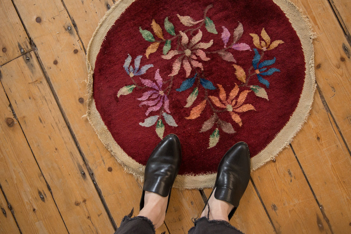 Round red rug with floral patterns on a wooden floor, with black shoes at the bottom.