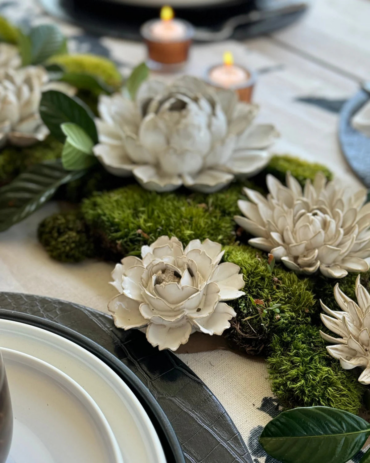 Decorative table setting with white ceramic flowers, green moss, and candles on a white tablecloth.