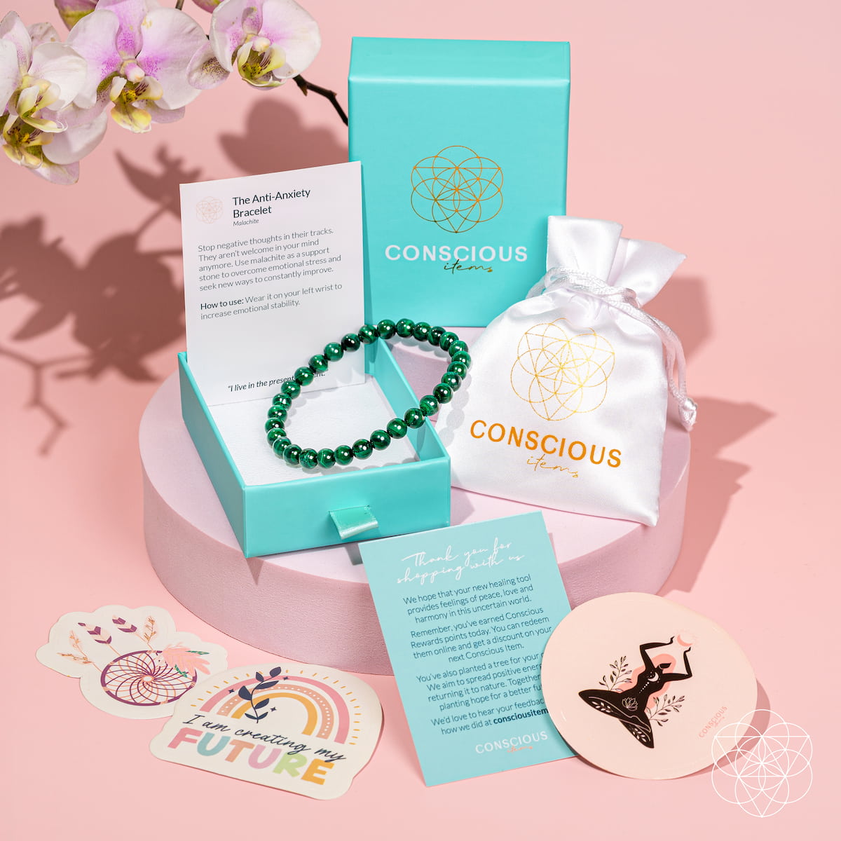 A Conscious Brand 'The Axiom Anxiety Bracelet' green beaded bracelet is presented in a turquoise jewelry box alongside its branded packaging, including a white drawstring pouch, cards, and decorative stickers on a pink background.