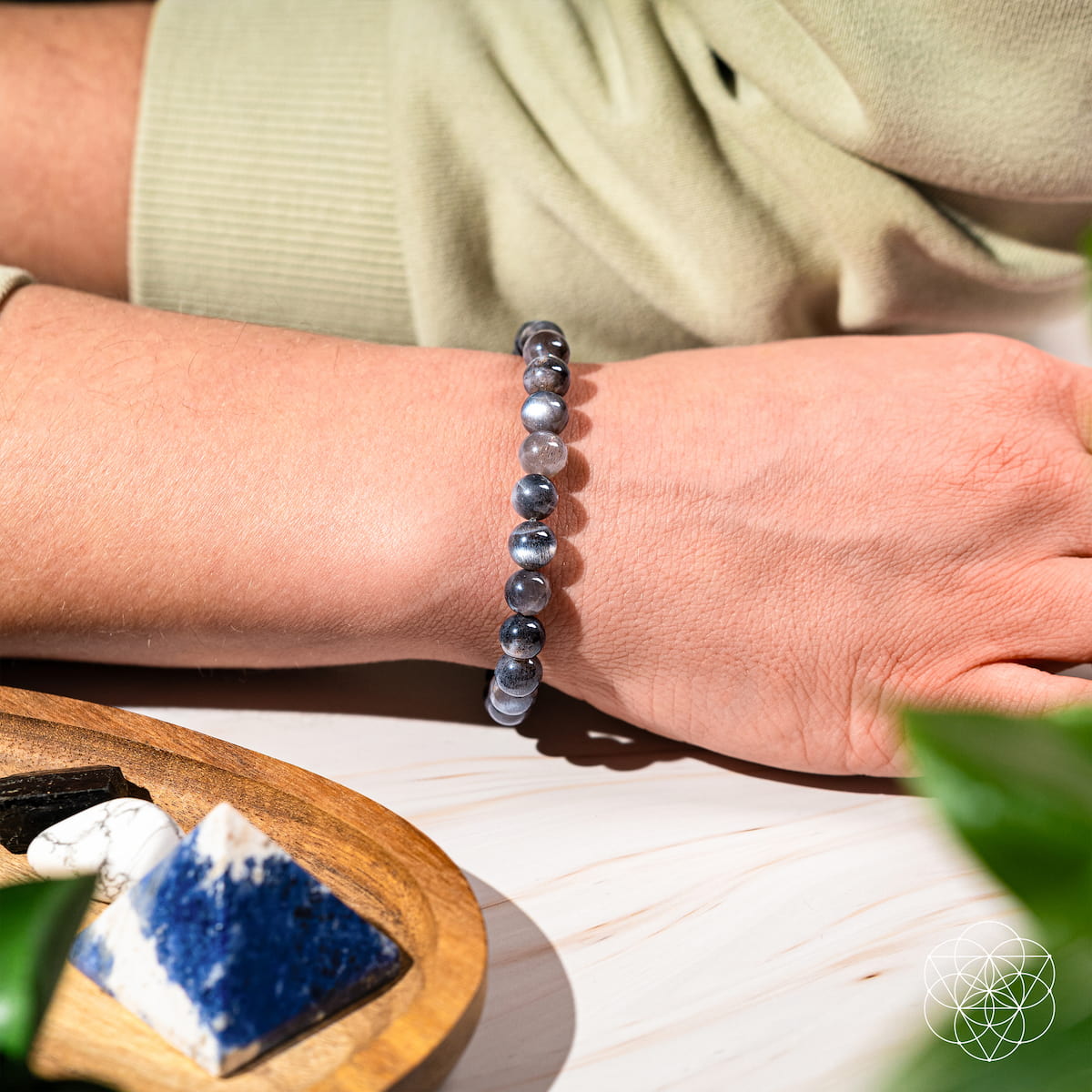 Person wearing a blue beaded bracelet on a light surface with a wooden tray and stones.