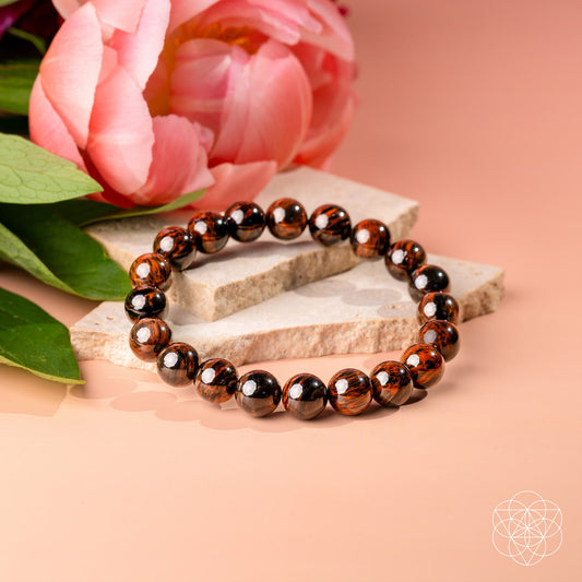 A beaded stretch bracelet made of glossy dark brown and black round beads, styled on stacked light stone slabs with a pink flower in the background on a peach-colored surface.