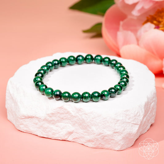 A green beaded bracelet made of polished oval beads, displayed on a white textured stone stand against a soft pink background with blurred pink flowers in the background.