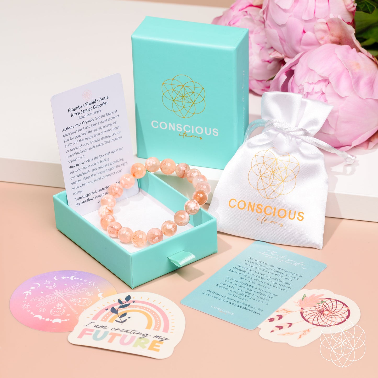 A Conscious Energy brand gemstone bracelet in a peachy-orange hue, displayed inside its mint green presentation box next to a white drawstring pouch, cards, and a pink flower arrangement in the background.