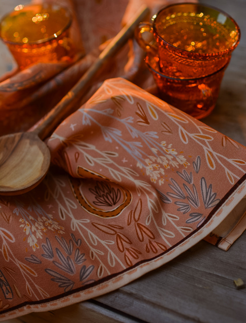 Open book with floral pattern on a wooden surface, accompanied by two amber-colored mugs and a wooden spoon.
