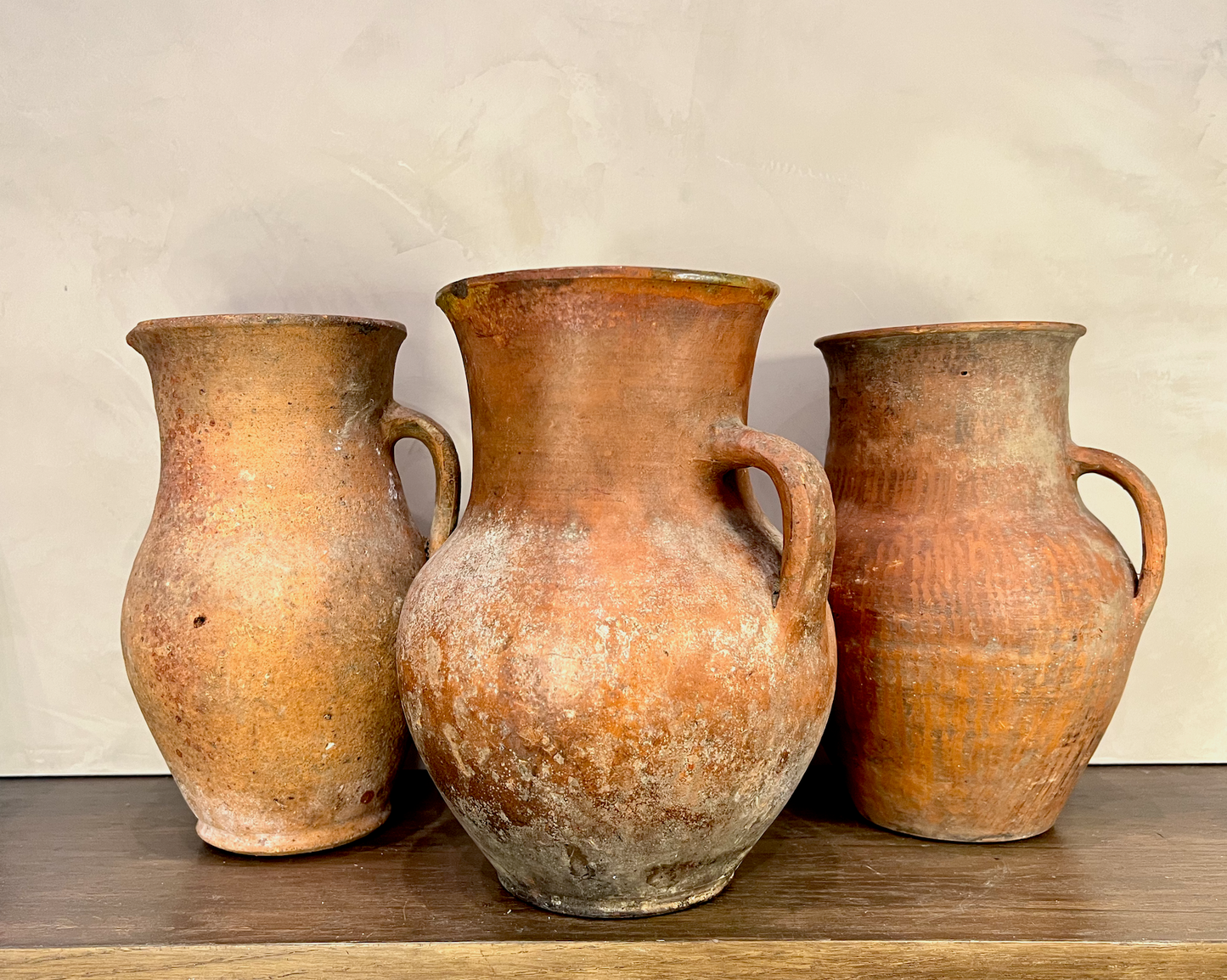 Three rustic terracotta clay jugs with handles, displayed on a wooden surface against a textured beige wall.