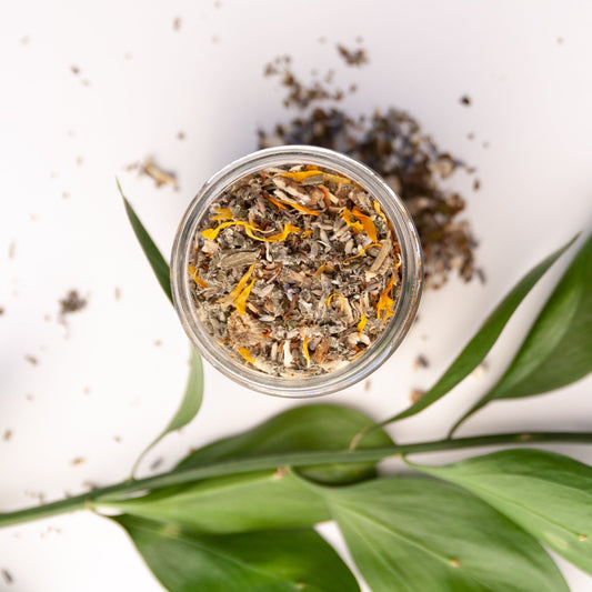 Glass jar filled with dried herbs and flowers on a white surface with green leaves.