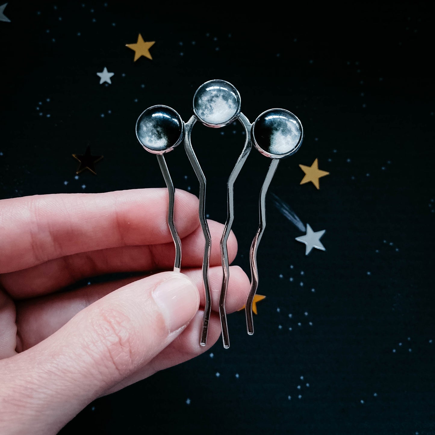 Hand holding four metallic hairpins with star and moon designs against a dark background with star illustrations.