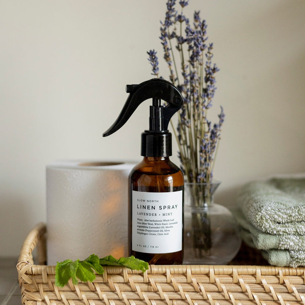 Brown linen spray bottle with black sprayer on a woven tray with lavender and a candle.