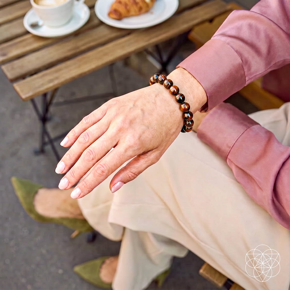 A close-up lifestyle shot of a person's hand wearing a brown and black beaded bracelet, seated at a wooden cafe table with a coffee cup and croissant in the background.