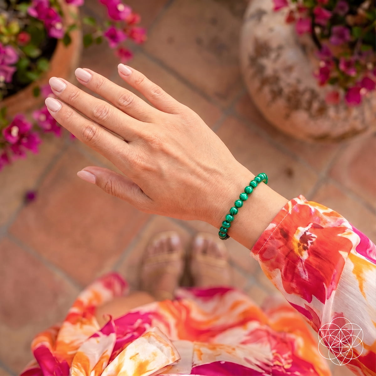 A close-up of a person's hand wearing a green beaded bracelet, with a colorful floral dress and terracotta tile flooring in the background.