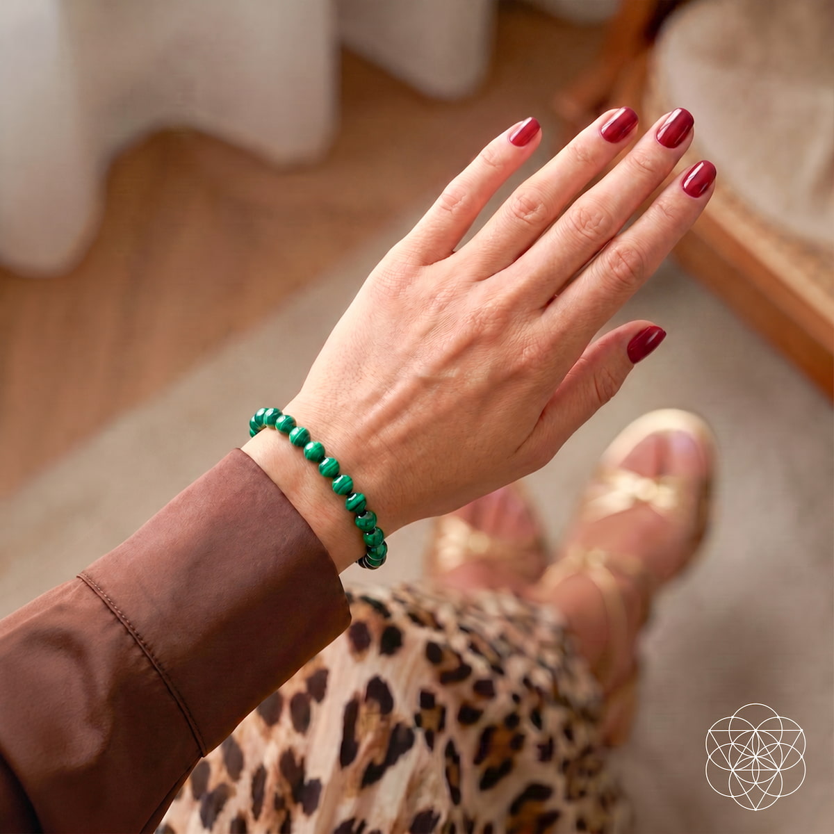 A close-up lifestyle shot of a person's hand wearing a green beaded bracelet, with red manicured nails, a brown sleeve, and a leopard print skirt visible in a softly blurred indoor setting.