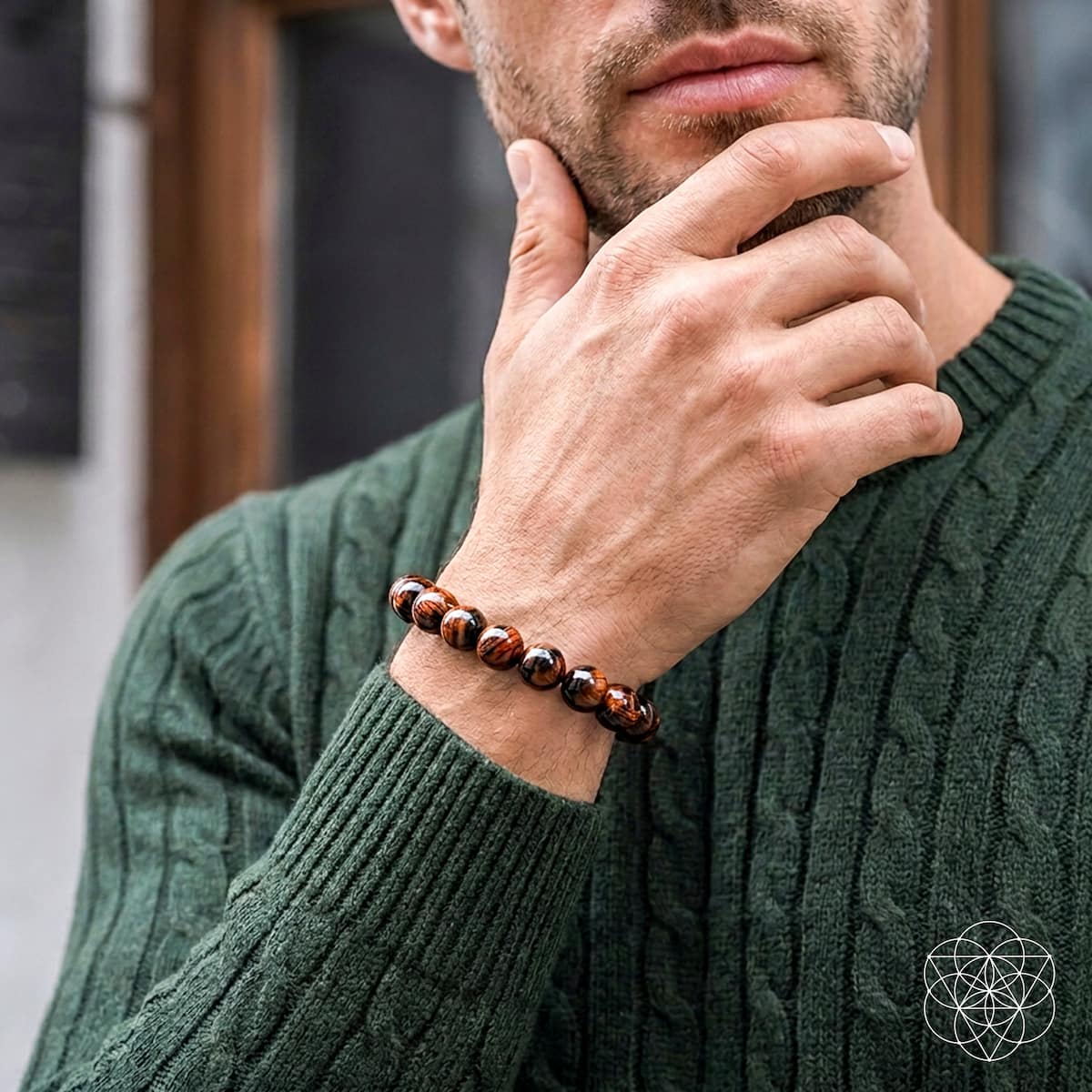 A close-up of a man wearing a dark green cable-knit sweater and a beaded bracelet on his wrist. The bracelet features glossy brown and black tiger's eye beads.