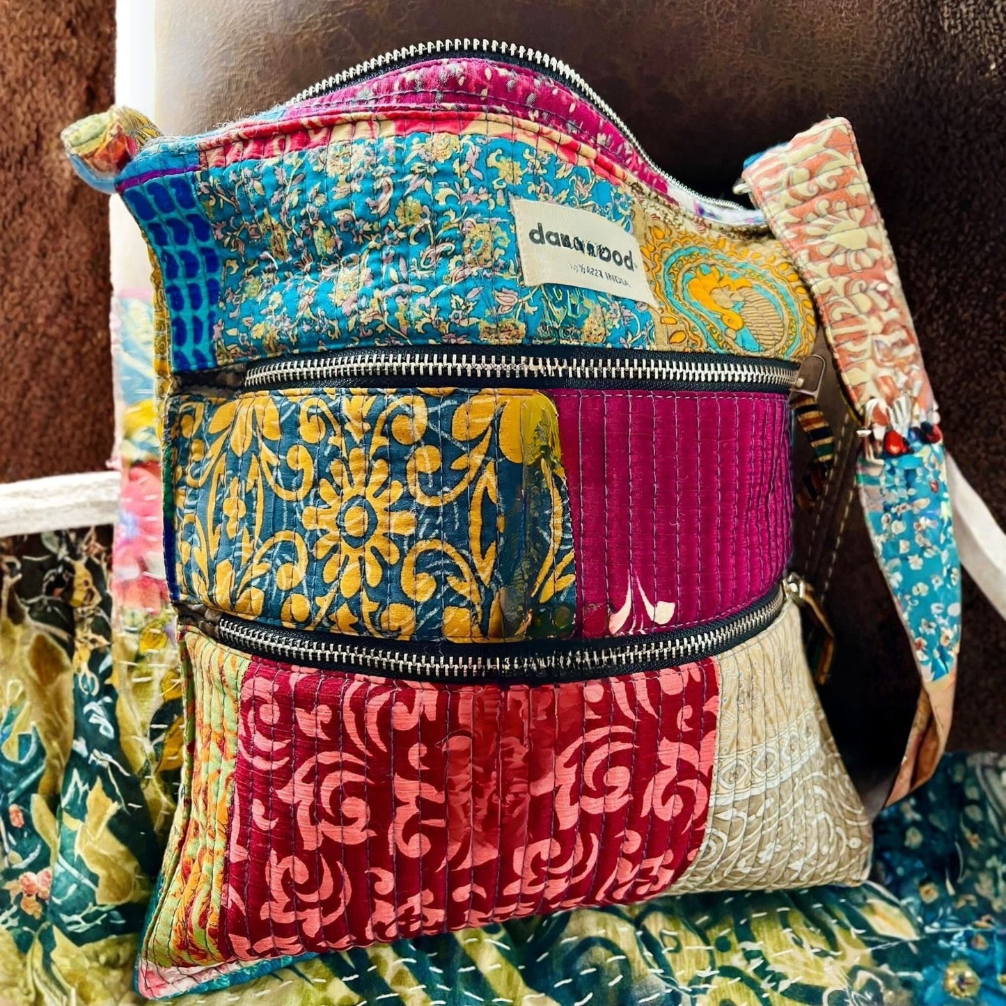 A colorful patchwork quilted crossbody bag with multiple zippered pockets, featuring vibrant patterns in blue, yellow, pink, and red. The brand label 'dakkahood' is visible on the front.