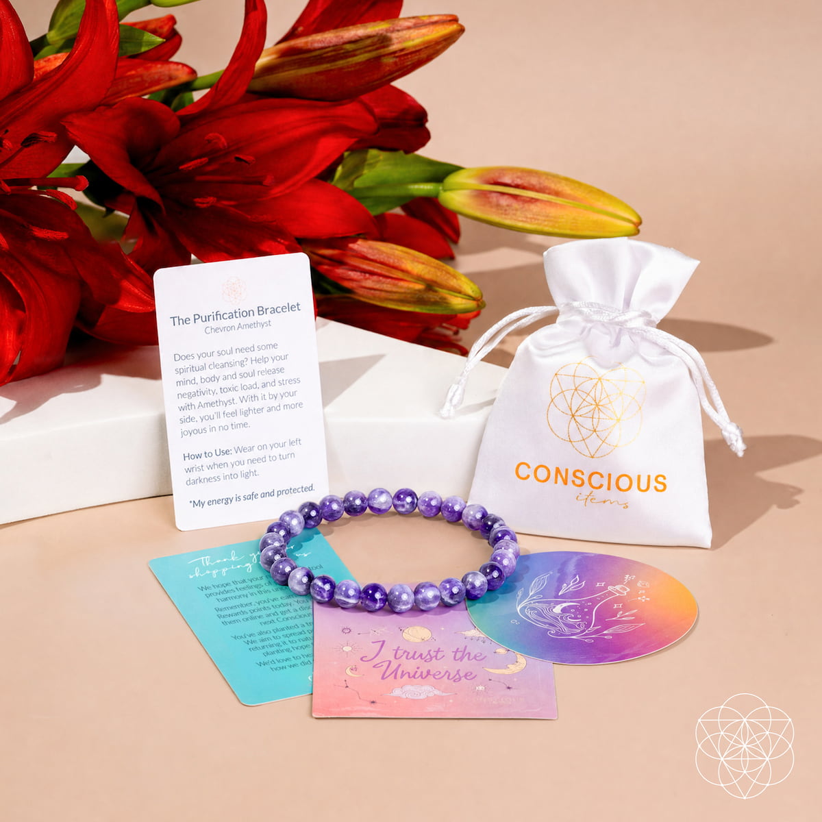 A purple amethyst bead bracelet is displayed with its gift set accessories, including a white drawstring pouch with a gold logo, a white instruction card, a teal affirmation card, and an iridescent oval card, arranged on a beige surface with red flowers in the background.