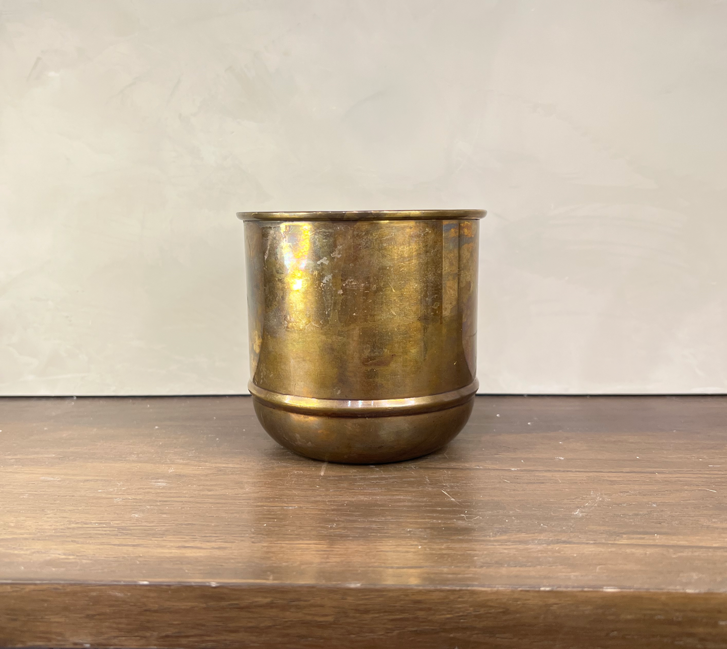 A vintage brass cup or tumbler with a rounded base, sitting on a wooden surface against a neutral light gray wall.