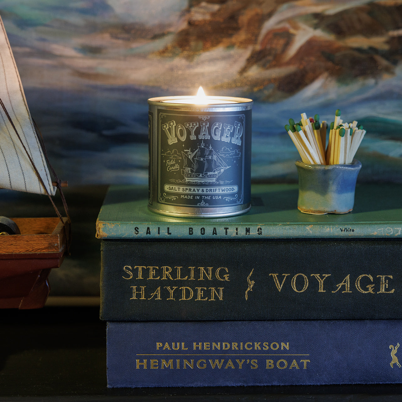 Candle in a 'Voyage' tin on top of books with a painting in the background