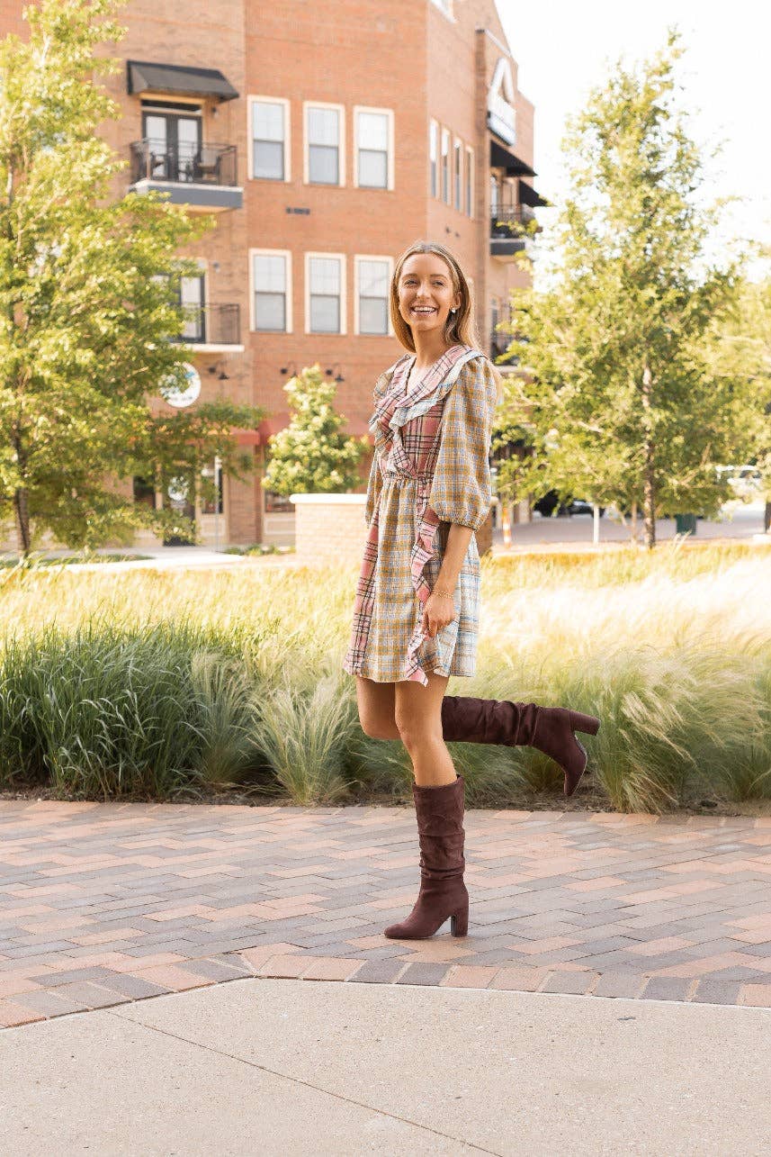 Woman in plaid dress and brown knee-high boots standing on a sidewalk with trees and buildings in the background.