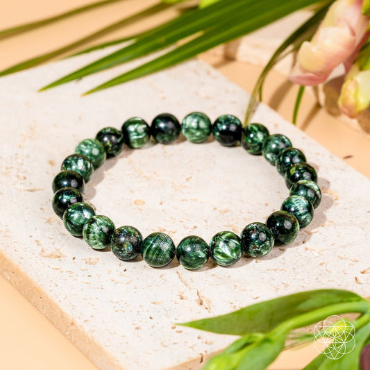 A beaded stretch bracelet made of polished dark green stone beads with natural marbled patterns, displayed on a textured beige stone slab with green leaves and soft pink flowers in the background.