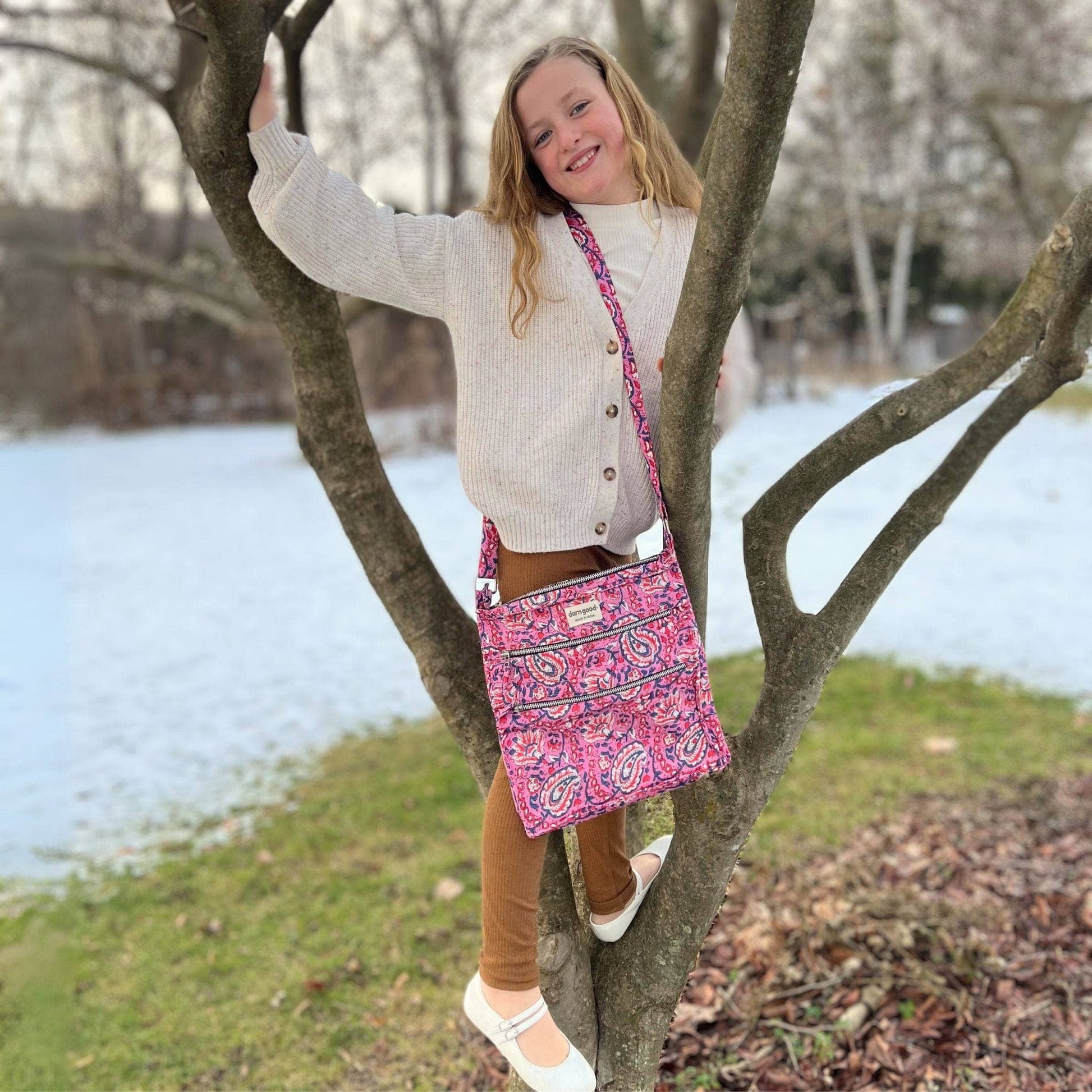 Child with a pink patterned bag standing in a tree by a body of water.