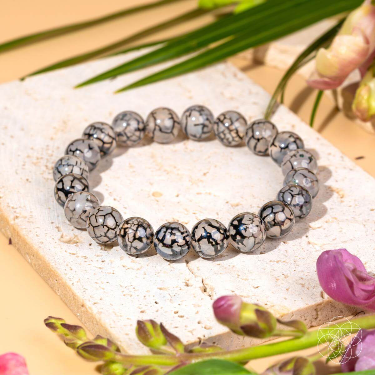A beaded stretch bracelet made of polished round beads with a black and white crackled pattern, displayed on a light stone slab with green leaves and pink flowers.
