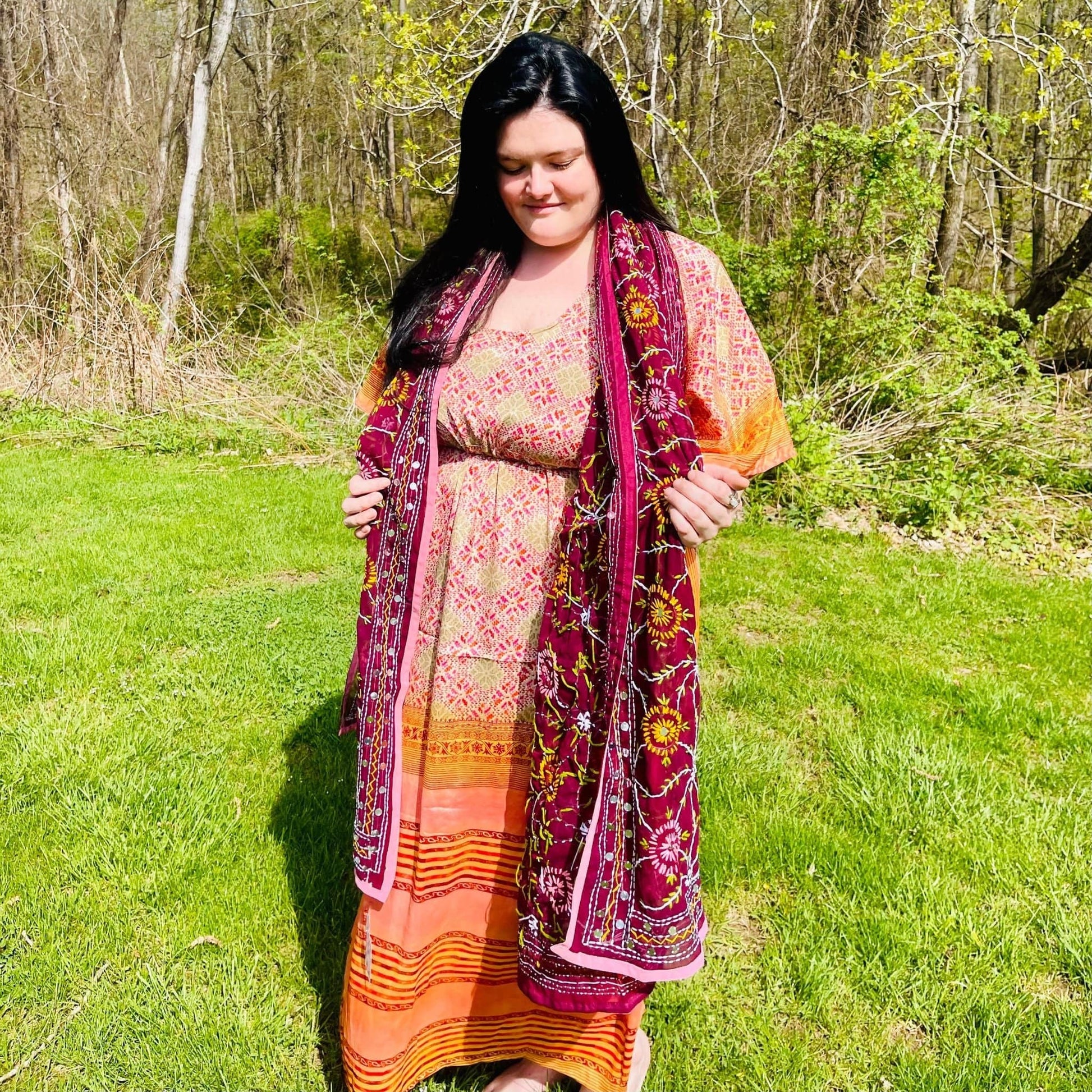 Woman in a colorful dress and shawl standing in a grassy area with trees.