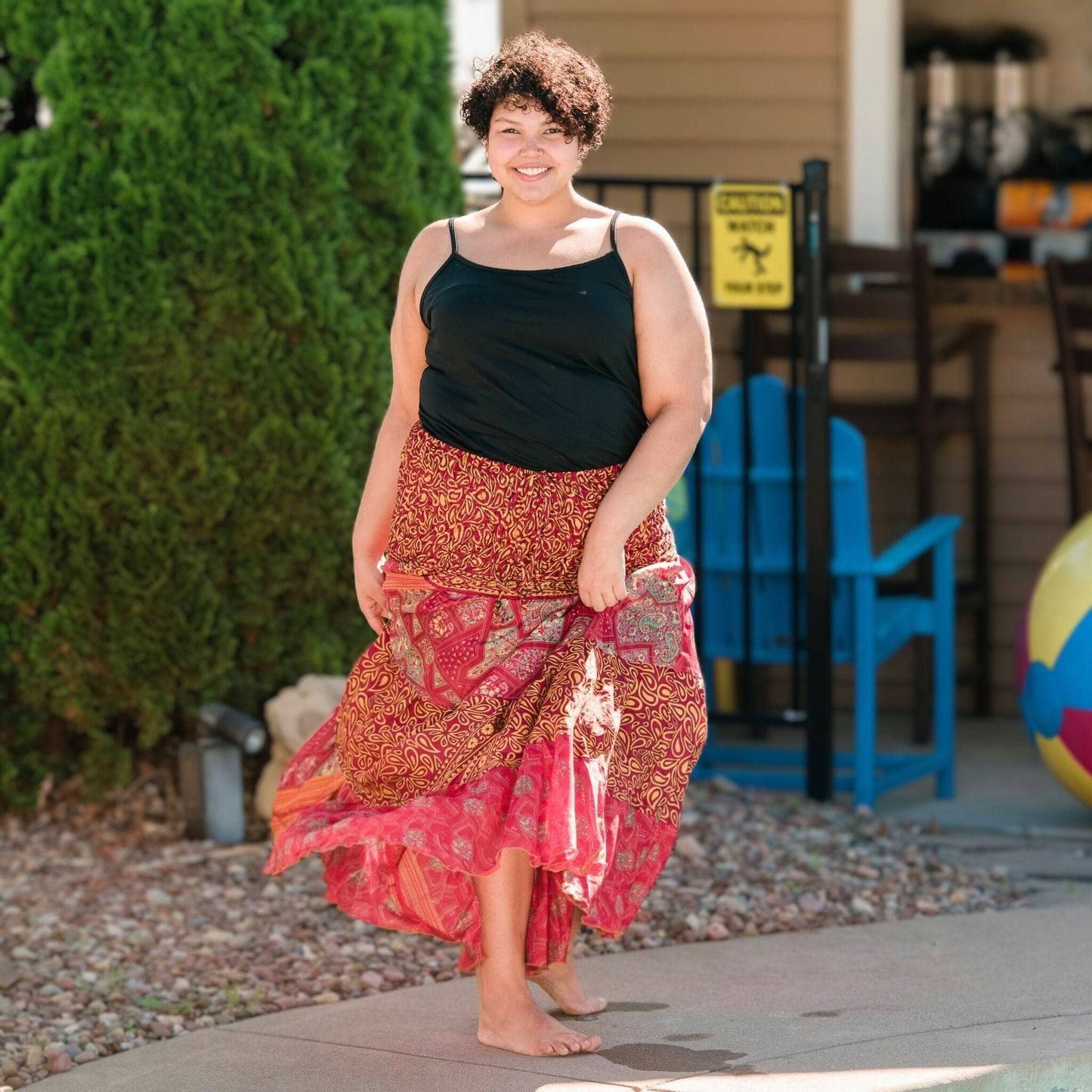 Woman in a black top and red patterned skirt standing outdoors with greenery and a chair in the background.