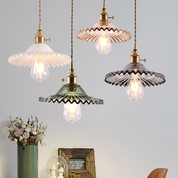 Four vintage-style pendant lights with scalloped glass shades in white, green, and smoky purple, hanging from brass cords against a light gray wall above a table with flowers and decor.
