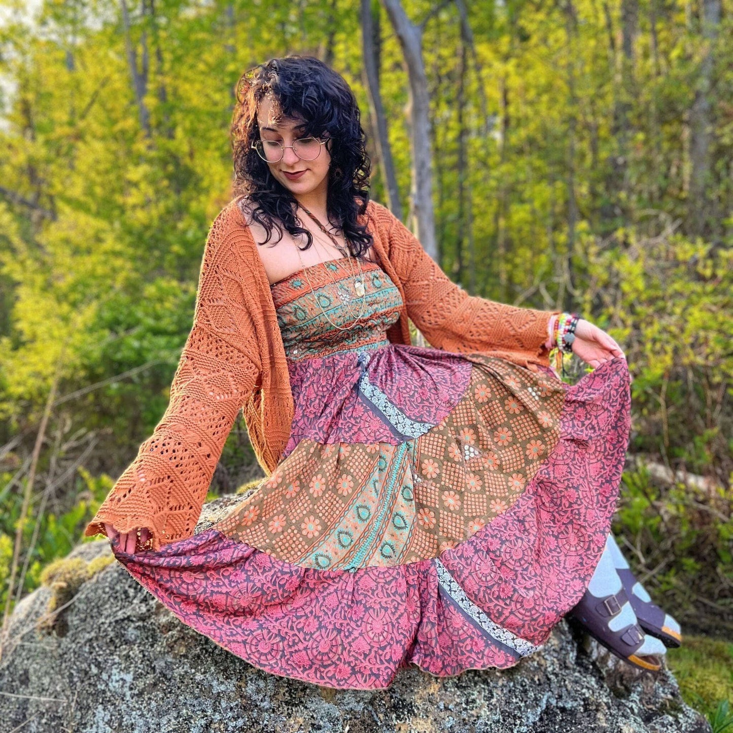 Woman in a colorful dress and cardigan sitting on a rock in a forest