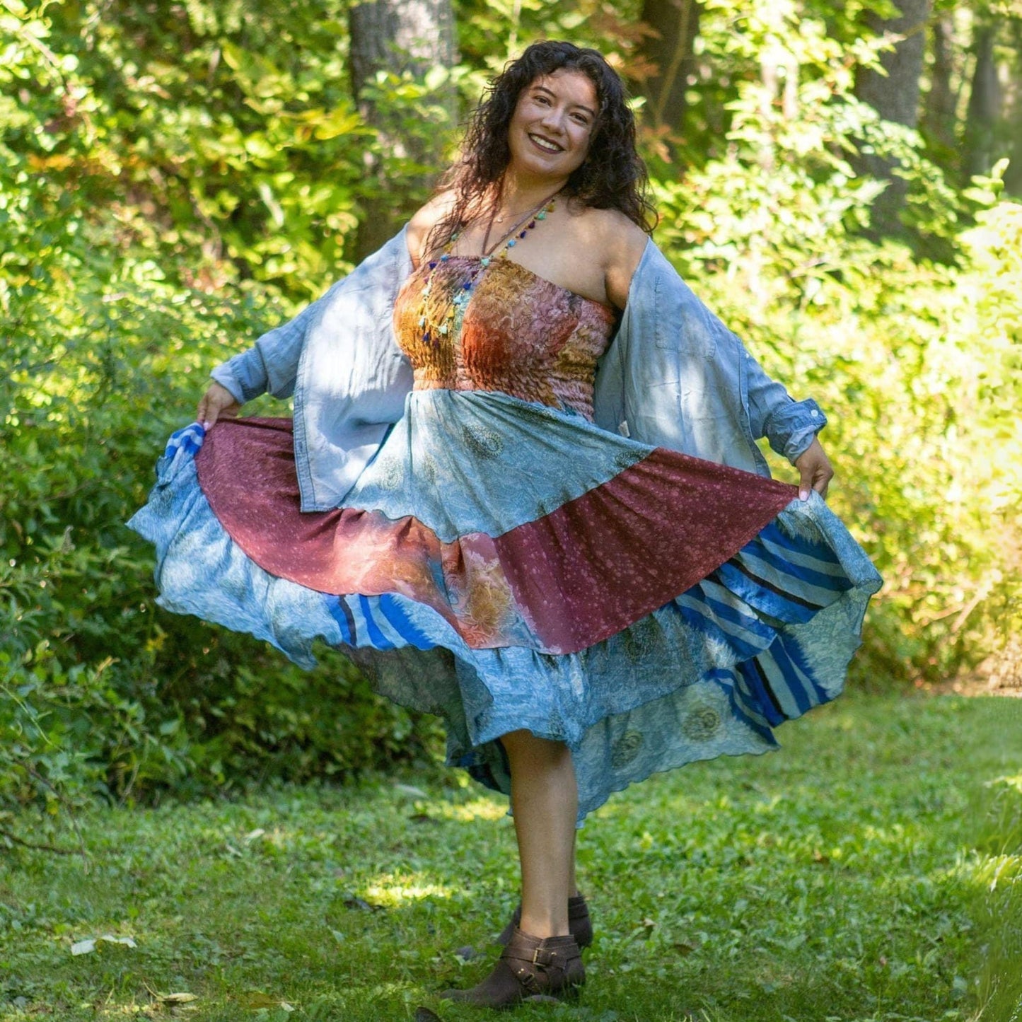 Woman in a colorful dress with a patterned top and flowing skirt standing in a grassy area with trees.