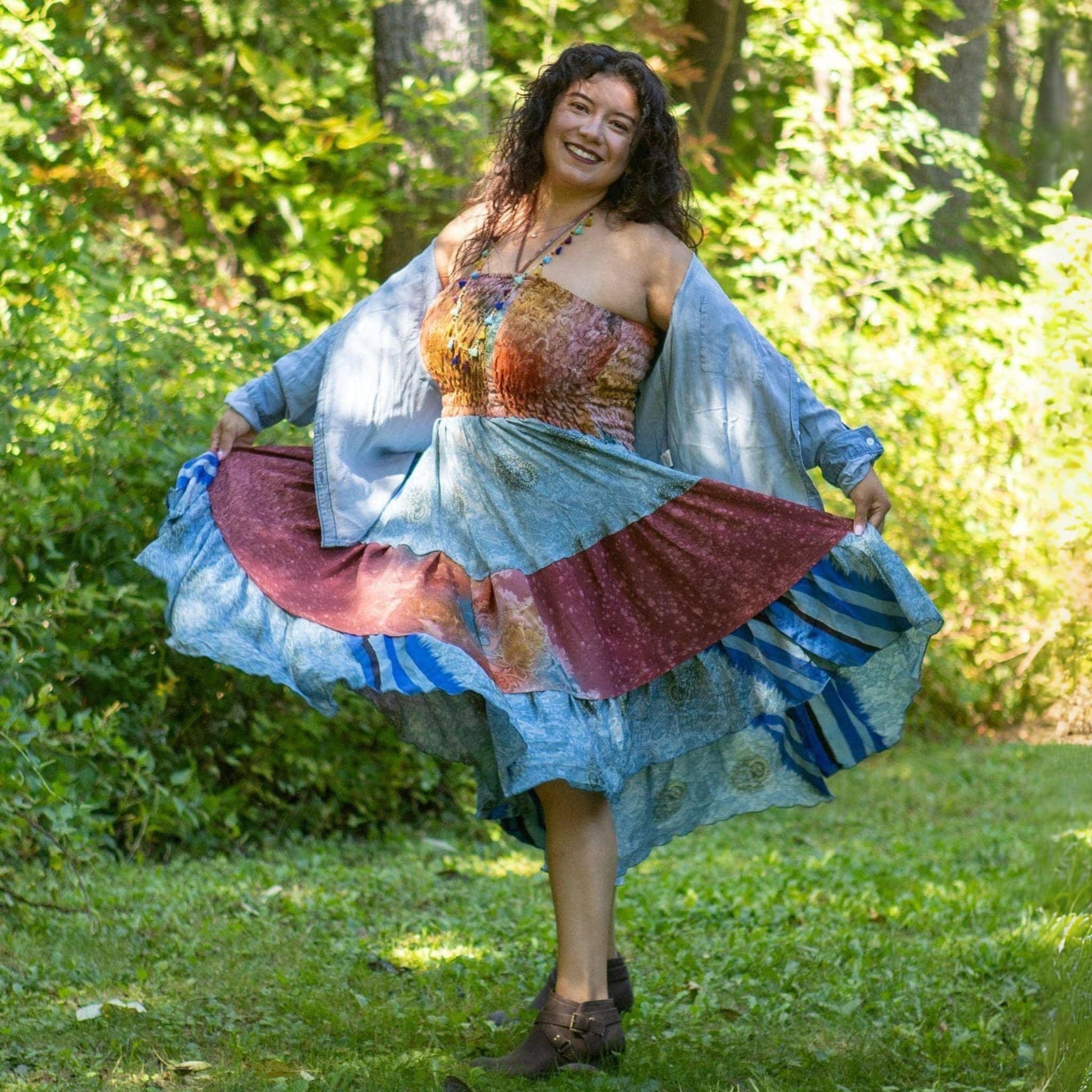 Woman in a colorful dress with a patterned top and flowing skirt standing in a grassy area with trees.