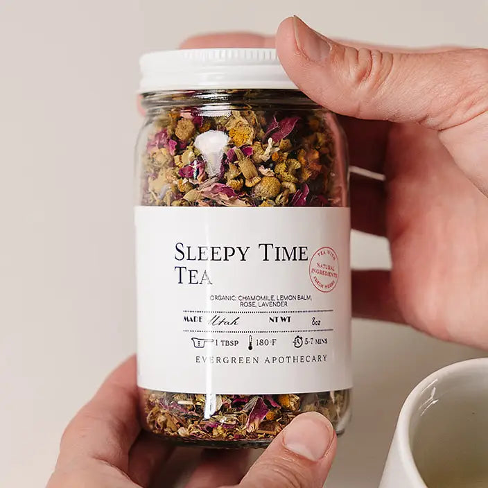 A hand holds a glass jar of Sleepy Time Tea by Evergreen Apothecary, showing the dried herbal blend inside, with a white mug partially visible in the foreground.