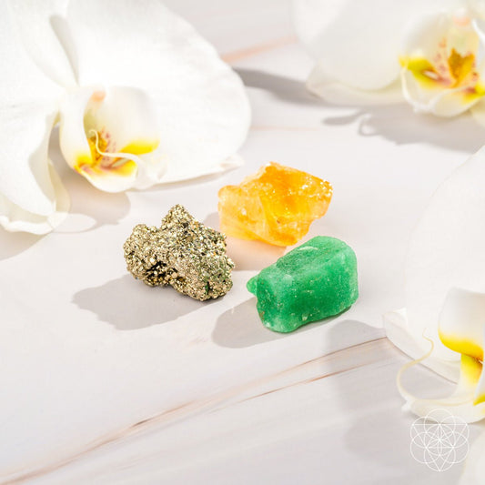 Three raw, unpolished crystals in green, yellow, and metallic gold are arranged on a white surface decorated with white orchid flowers.