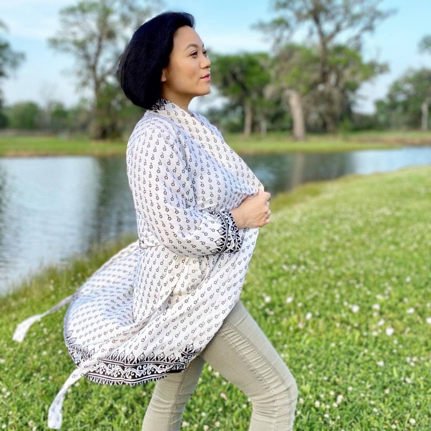 Woman in a patterned white and black outfit standing in a grassy area with a body of water in the background.