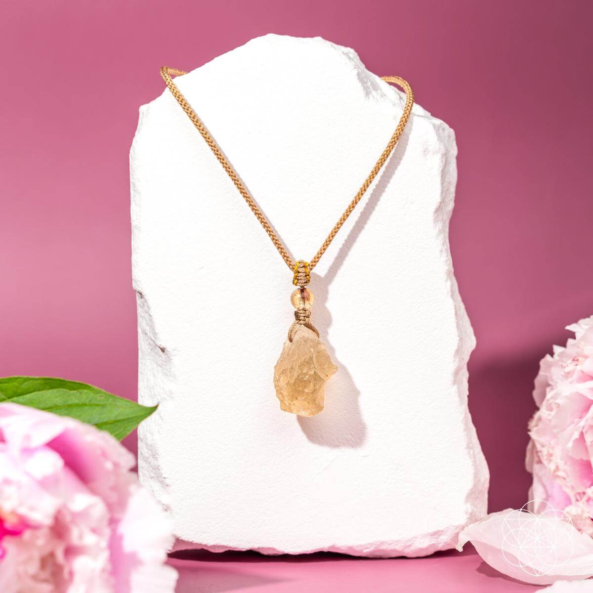 A gold chain necklace with a raw, irregular-shaped yellow crystal pendant, displayed on a white stone block against a pink background with pink flowers in the foreground.