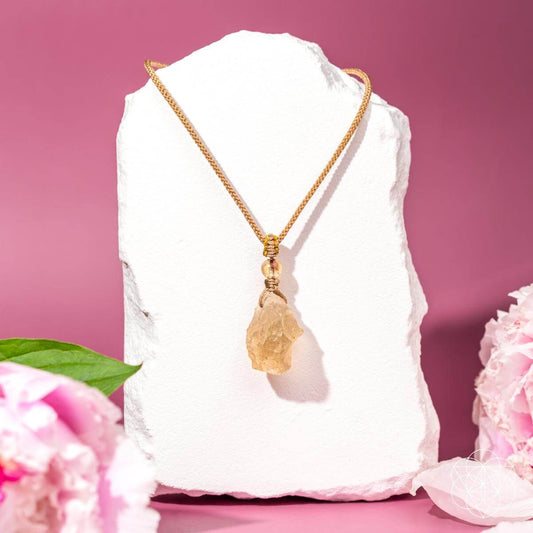 A gold chain necklace with a raw, irregular-shaped yellow crystal pendant, displayed on a white stone block against a pink background with pink flowers in the foreground.